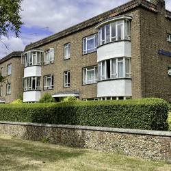 An image of a three-story brick building with white windows, behind a short wall and hedge. 