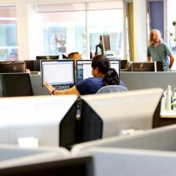 A woman is sat at a desk with her back to the camera, looking at a computer screen in an office. 
