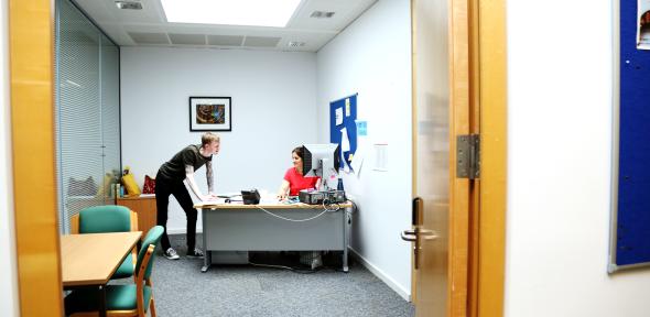 A woman sits at a deskin an office, and a man stands next to her. They appear to be talking.