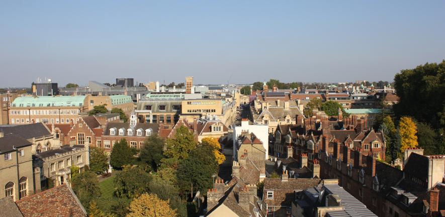 A photograph showing an aerial view over Cambridge city centre, taken from the Pitt Building. The sky is blue and between the rooftops there are green trees and grass. 